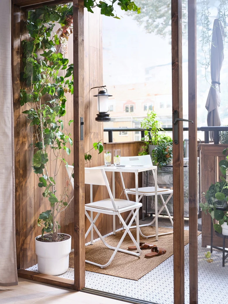 Malý balkon s květinami, bílým nábytkem a dřevěnými prvky.