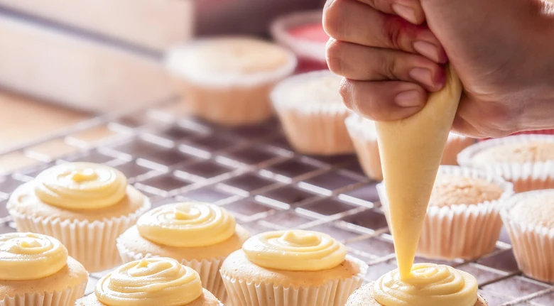 Ruka natíruje vanilkovou krémem šlehačku na muffiny.