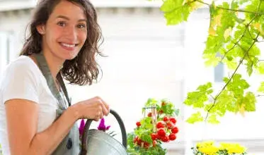 Žena v zahradnických kalhotách zalévá květiny na balkóně.